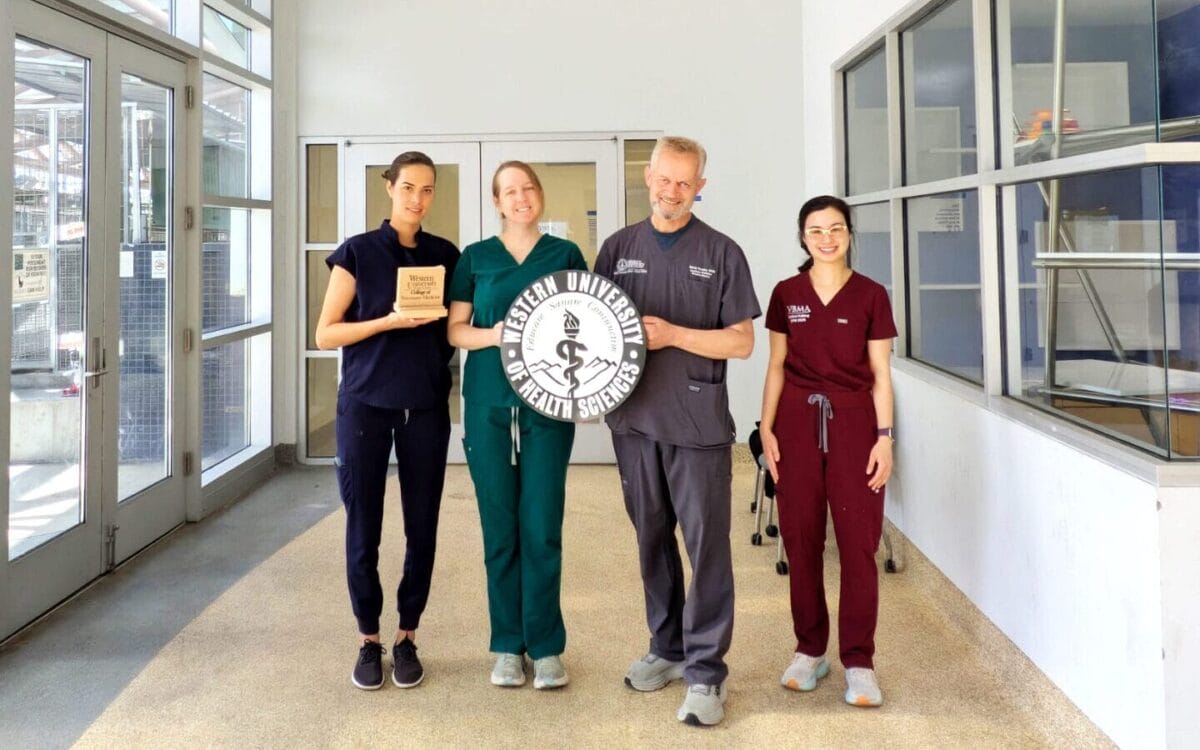 Four people in scrubs stand under an "Animal Clinic" sign in a hallway; two hold a plaque and a circular Western University College of Veterinary Medicine logo.