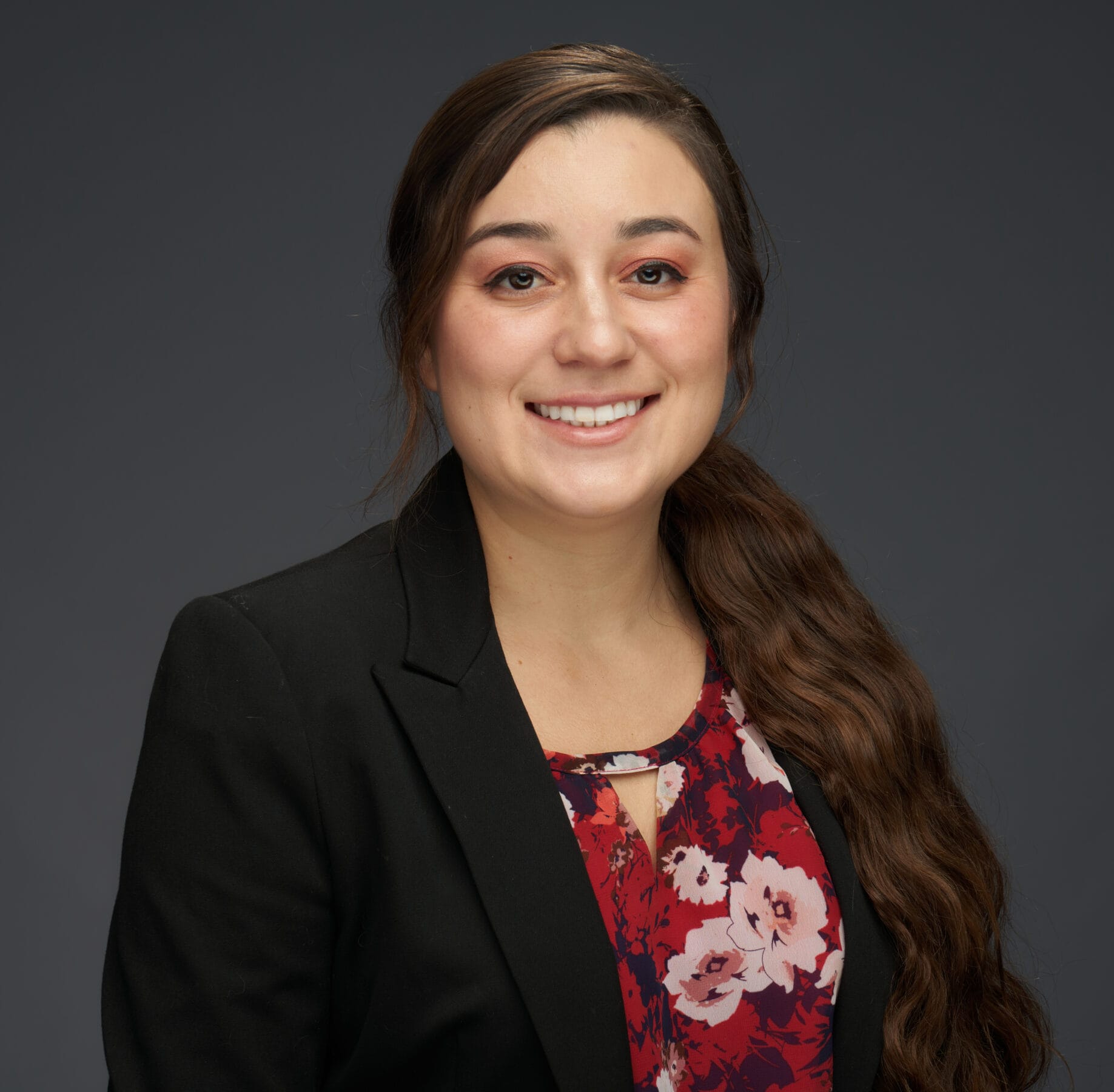 A woman with long brown hair in a side ponytail wears a black blazer over a red floral blouse and smiles at the camera against a plain dark background.
