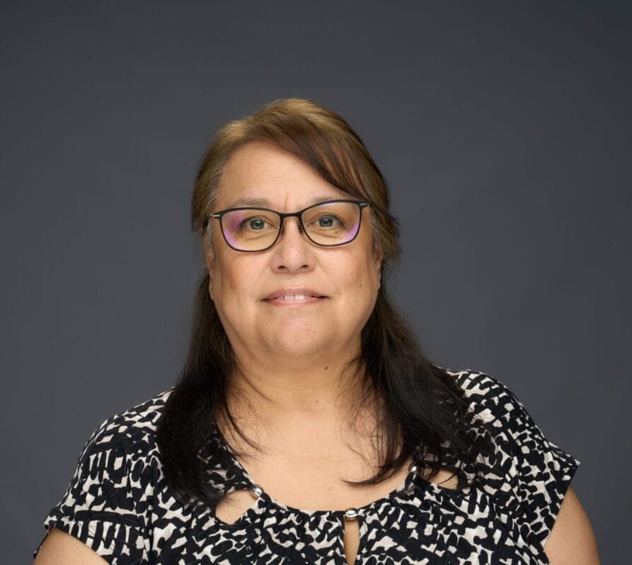 A woman with long brown hair and glasses, wearing a black and white patterned top, poses in front of a plain gray background.