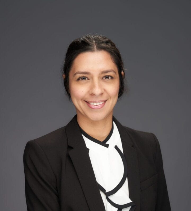 A woman wearing a black blazer and white blouse poses in front of a plain dark background, smiling at the camera.