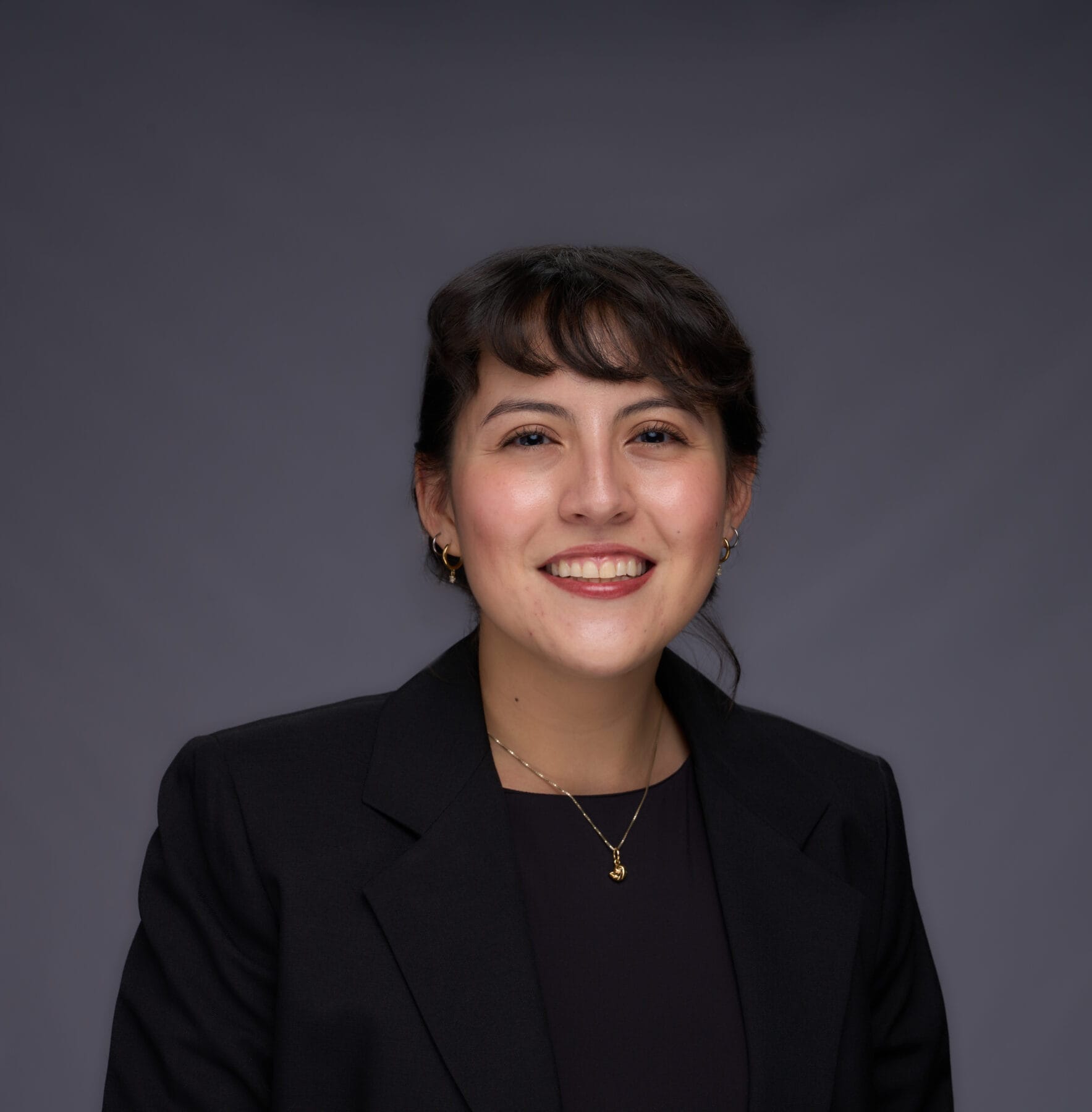 A woman with short dark hair wearing a black blazer and gold jewelry smiles at the camera against a plain dark background.