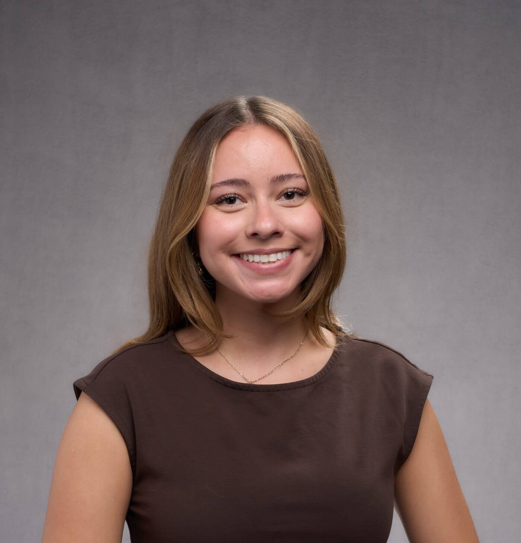 A young woman with straight light brown hair, wearing a dark brown short-sleeve top, smiles in front of a plain gray background.