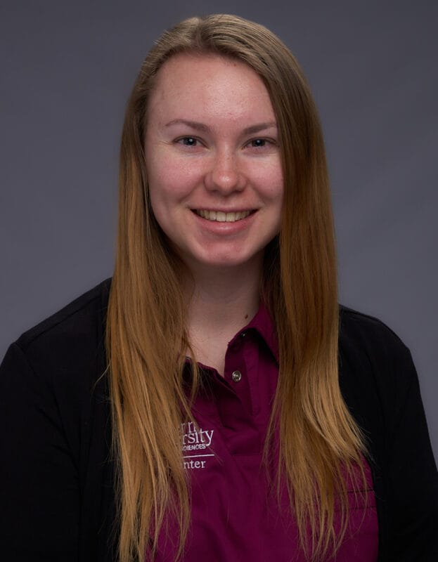 Woman with long blonde hair, wearing a maroon collared shirt and black sweater, smiling in front of a plain gray background.