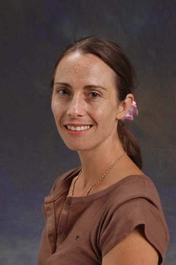 Woman with brown hair pulled back, wearing a brown top and gold necklace, smiling in front of a dark, gradient background.