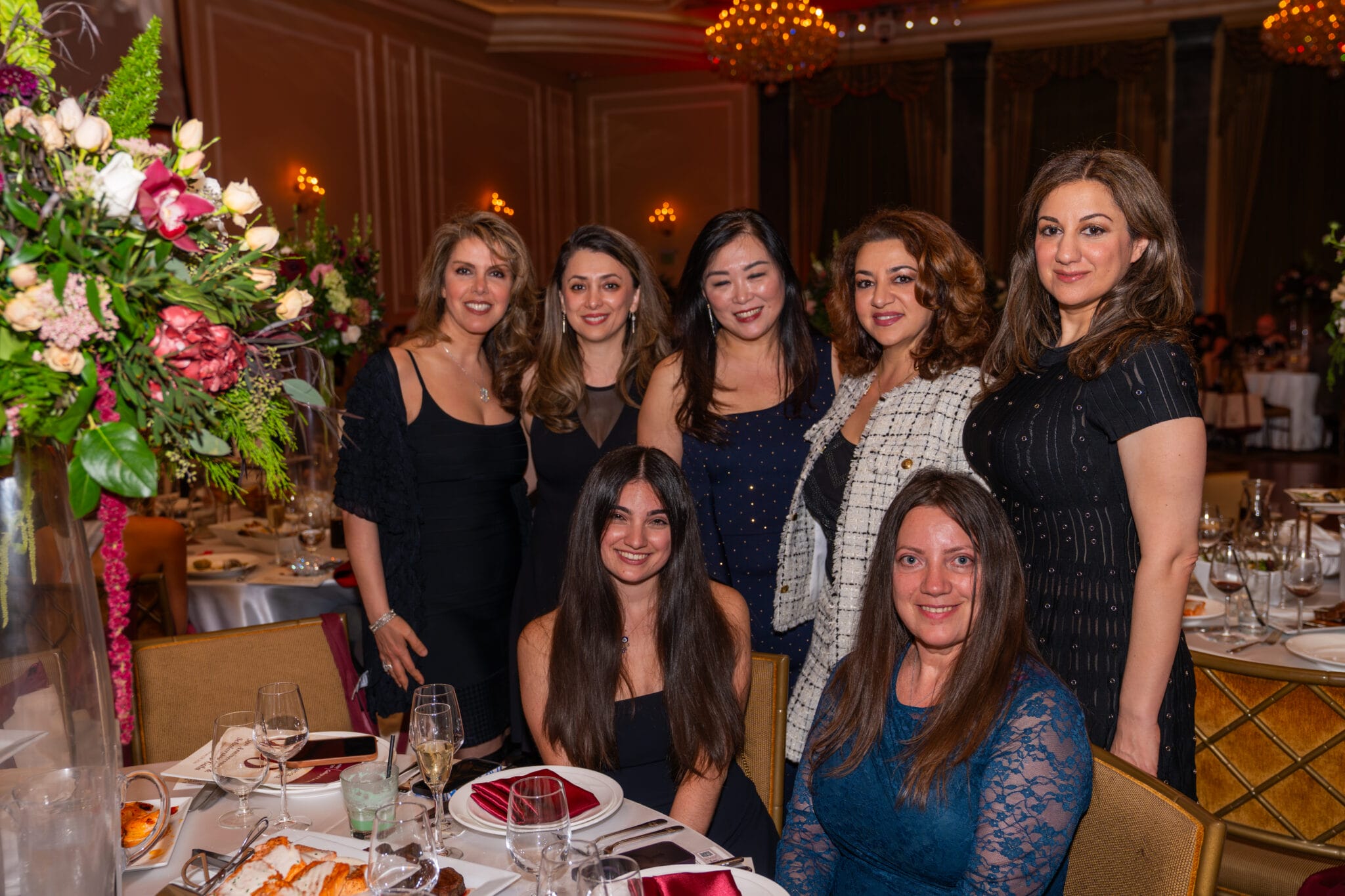 Seven women pose together at a formal event, with elegant table settings, floral arrangements, and chandeliers visible in the background.