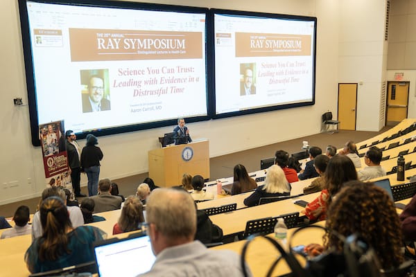 A speaker presents at the Ray Symposium in a lecture hall, with an audience seated and a presentation titled "Science You Can Trust: Leading with Evidence in a Distrustful Time" displayed on screens.