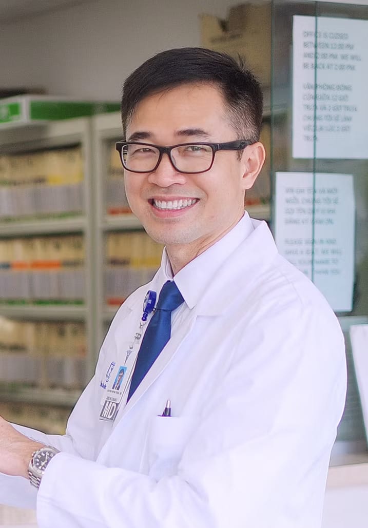 A man in a white lab coat and glasses stands in front of shelves filled with medical supplies, smiling at the camera.