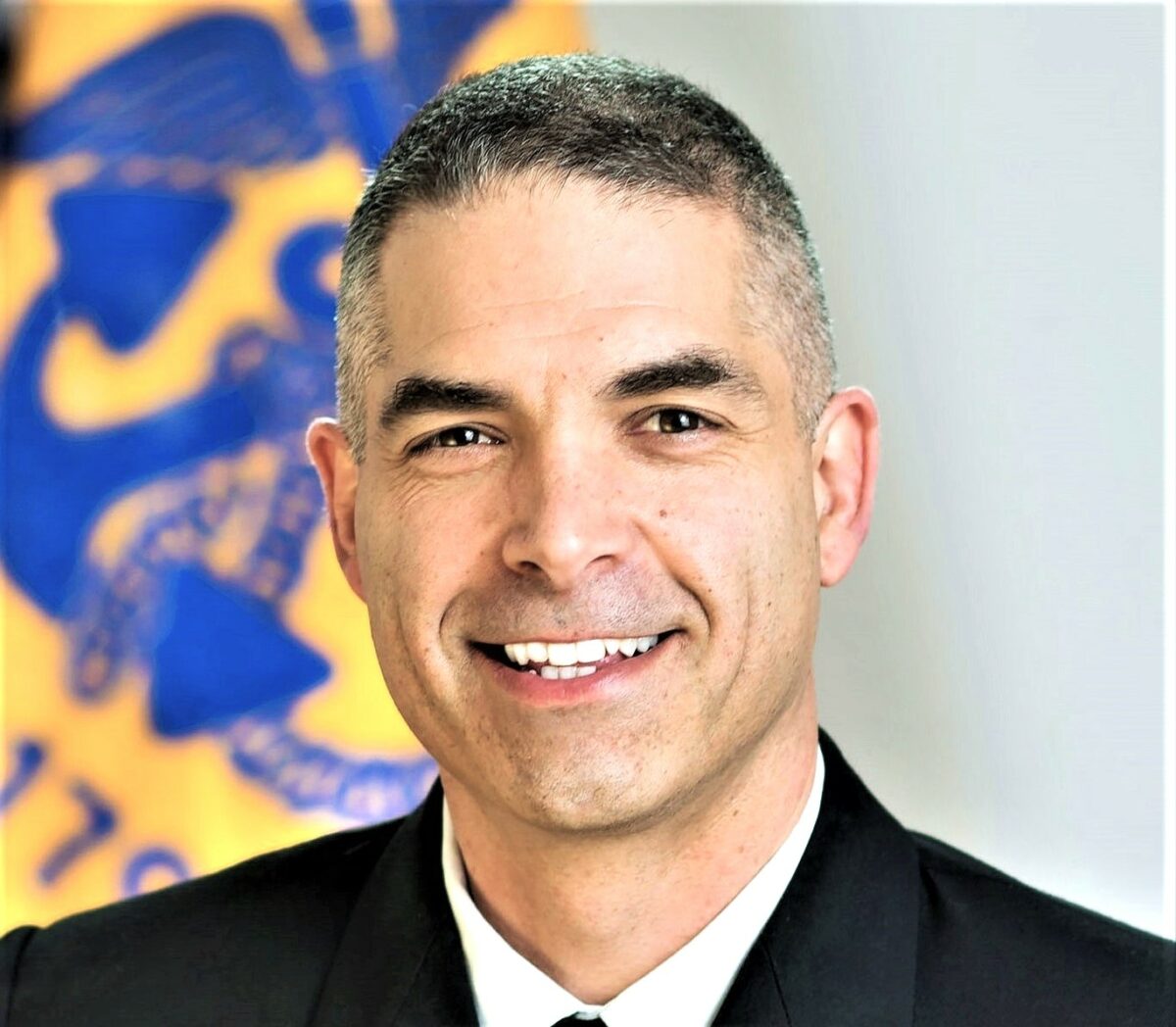 A man in uniform smiling in front of a flag.