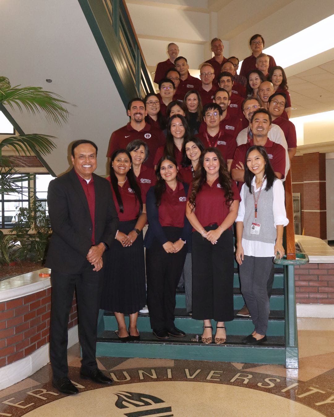 A group of people pose on a staircase inside a building, with most wearing matching maroon shirts and a man in a black suit standing in front.