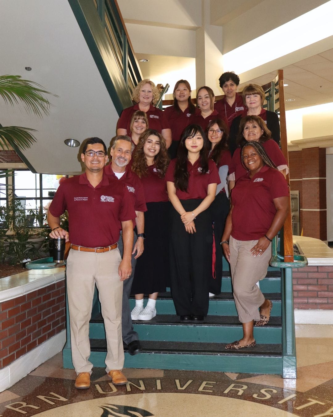 A group of thirteen people in maroon shirts and business attire pose together on a staircase inside a building with university signage visible.