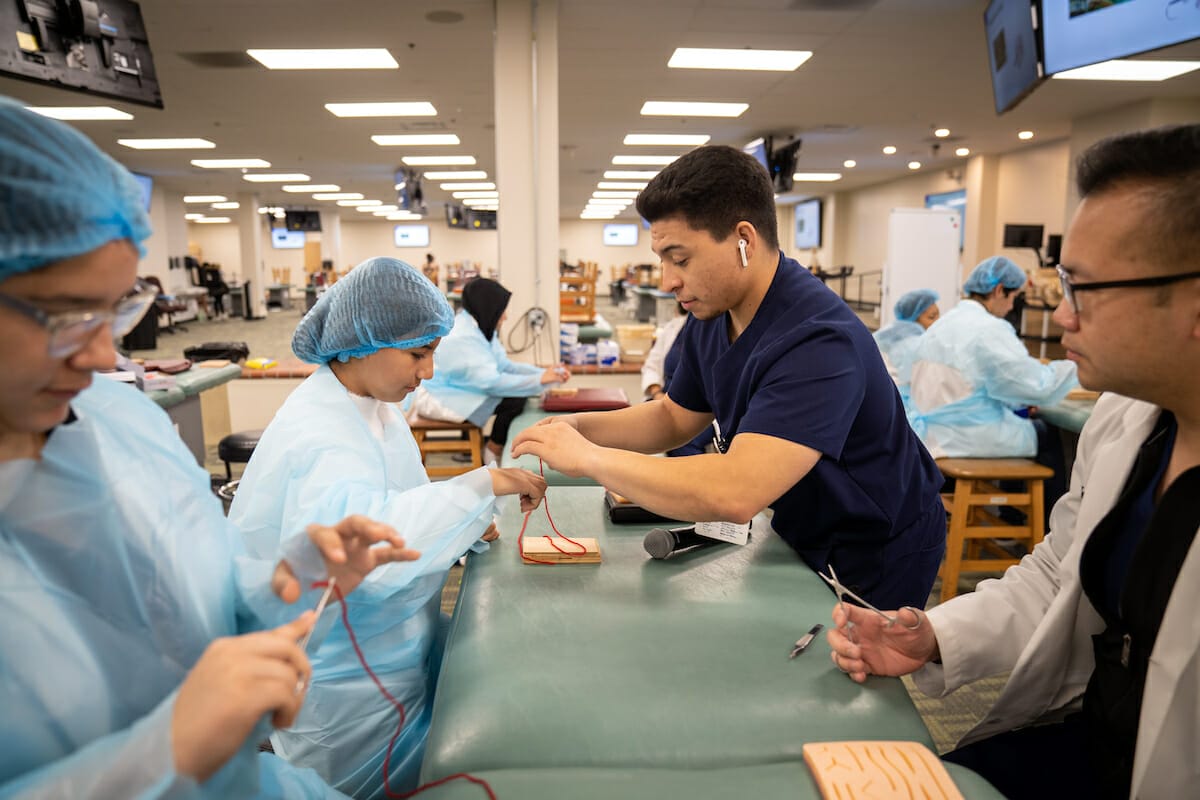 Medical students practice suturing on synthetic skin pads in a classroom, wearing blue gowns and hair covers, supervised by an instructor.