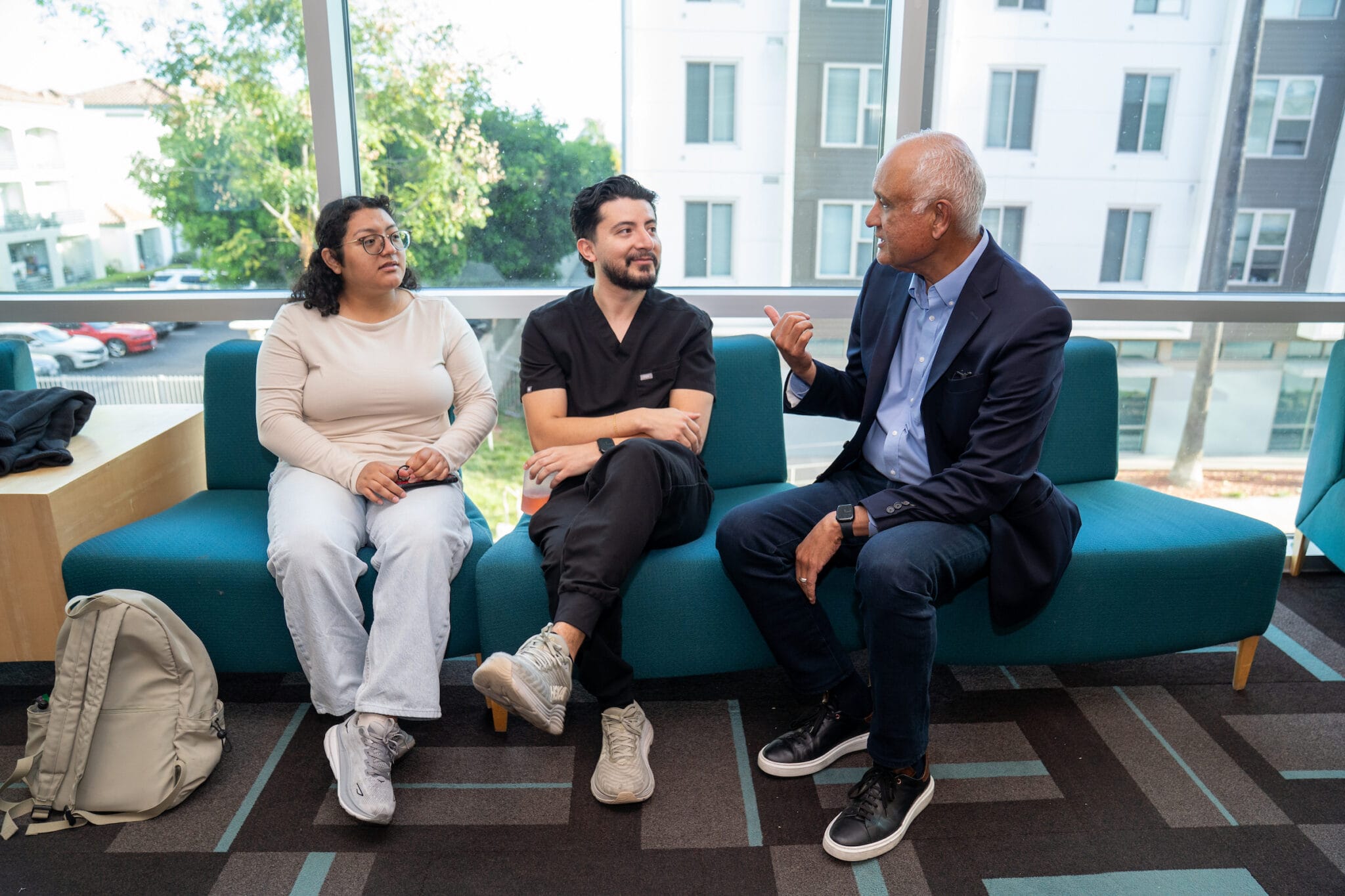 Three people sit on a teal couch in conversation near large windows; two are casually dressed, one is in business attire. A backpack rests beside them.