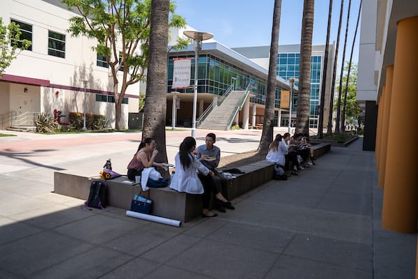 Students sit and talk on benches outside a modern campus building with palm trees and a staircase visible in the background.