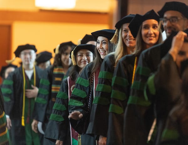 A group of graduates in academic regalia with green and black robes stand in line at a graduation ceremony.