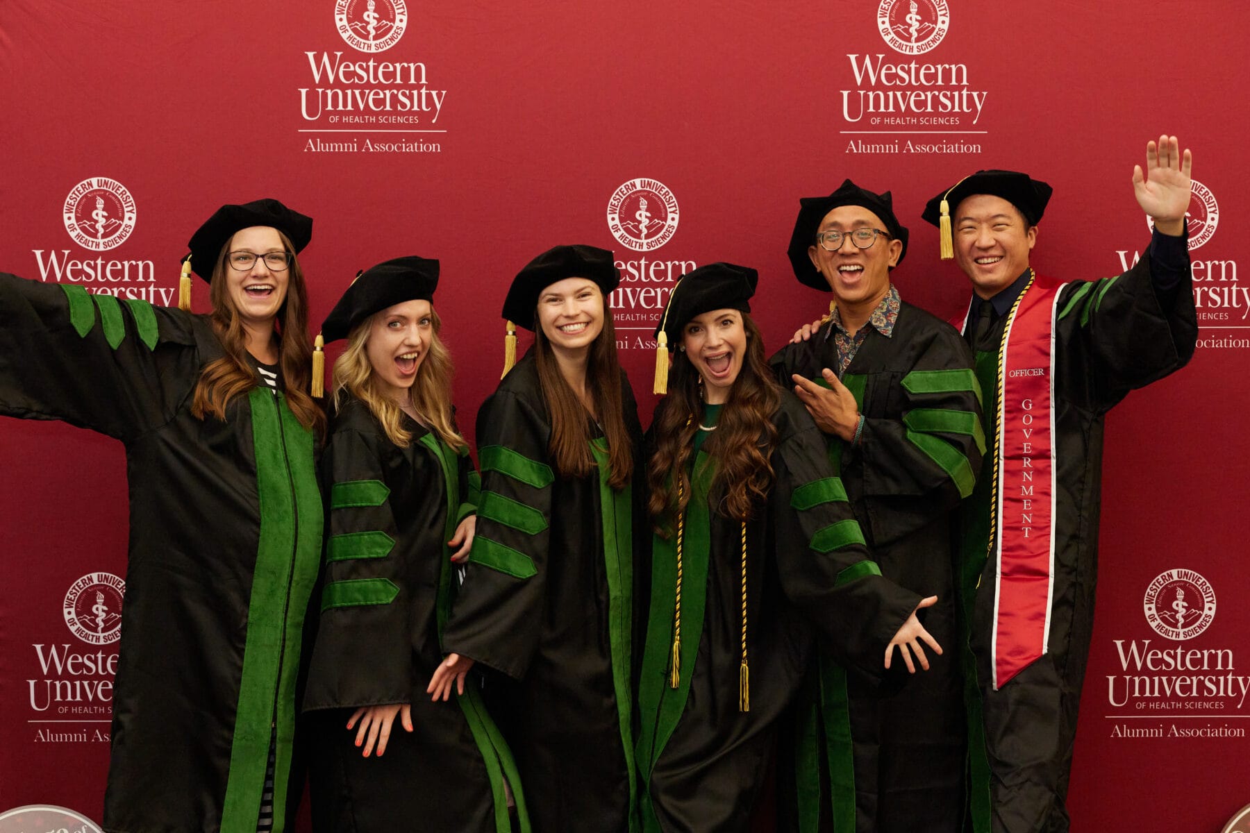 Six graduates in academic regalia pose and smile in front of a red Western University of Health Sciences Alumni Association backdrop.