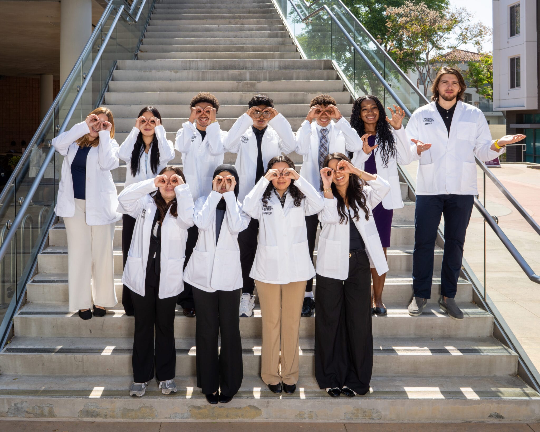 A group of ten people in white lab coats stand on steps outside, making hand gestures around their eyes and smiling at the camera.