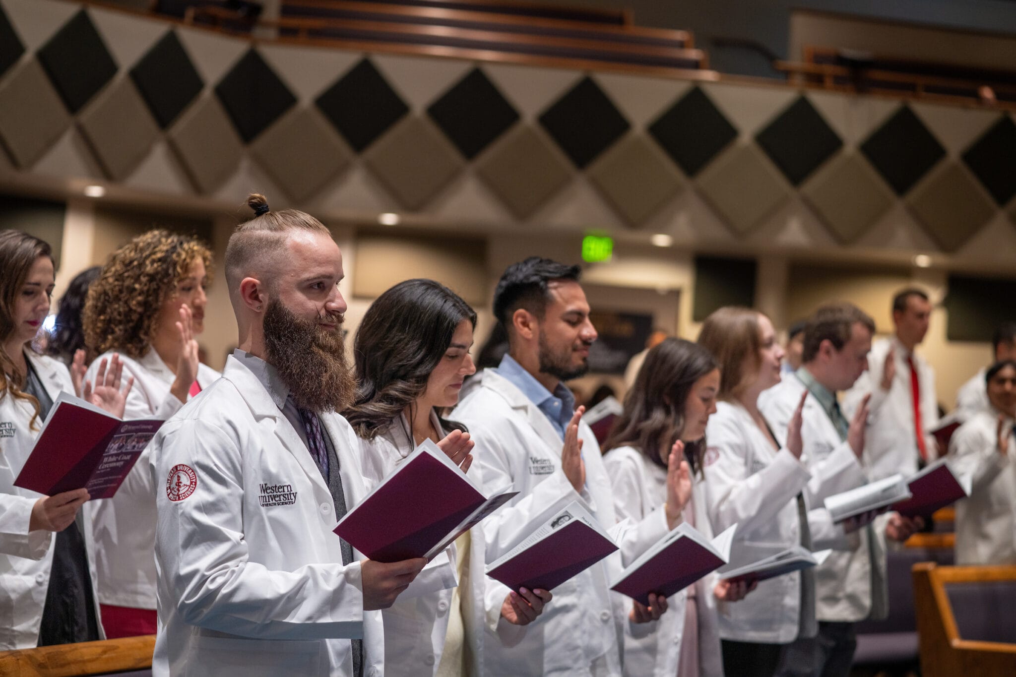 A group of students in white coats hold booklets and raise their right hands during a ceremony in an auditorium.