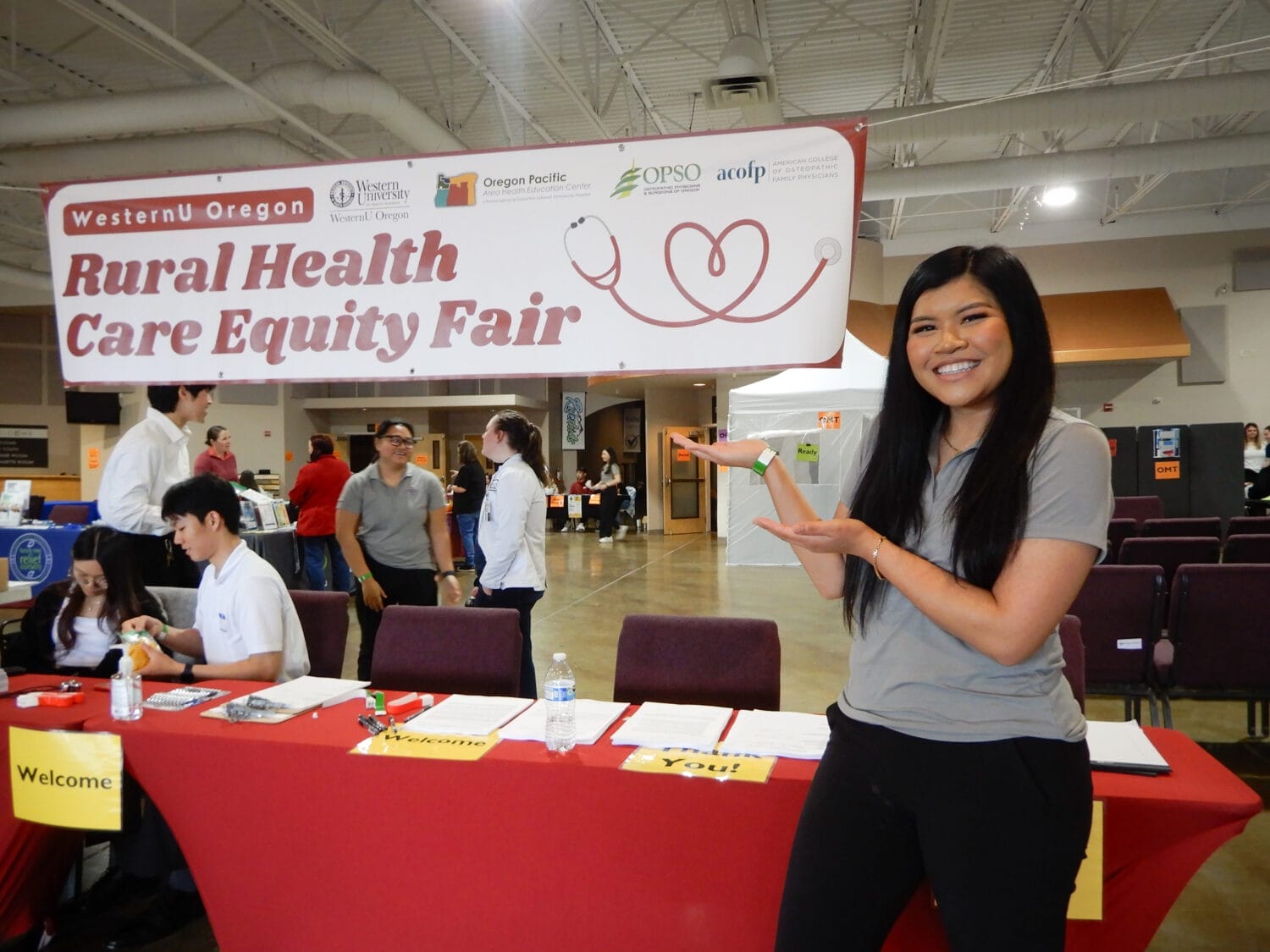 A woman stands smiling and gesturing toward a "Rural Health Care Equity Fair" sign at an indoor event with tables and attendees in the background.