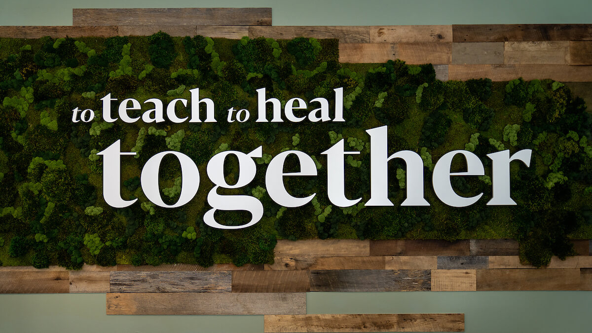 White text on a moss and wood wall reads: “to teach to heal together.”.