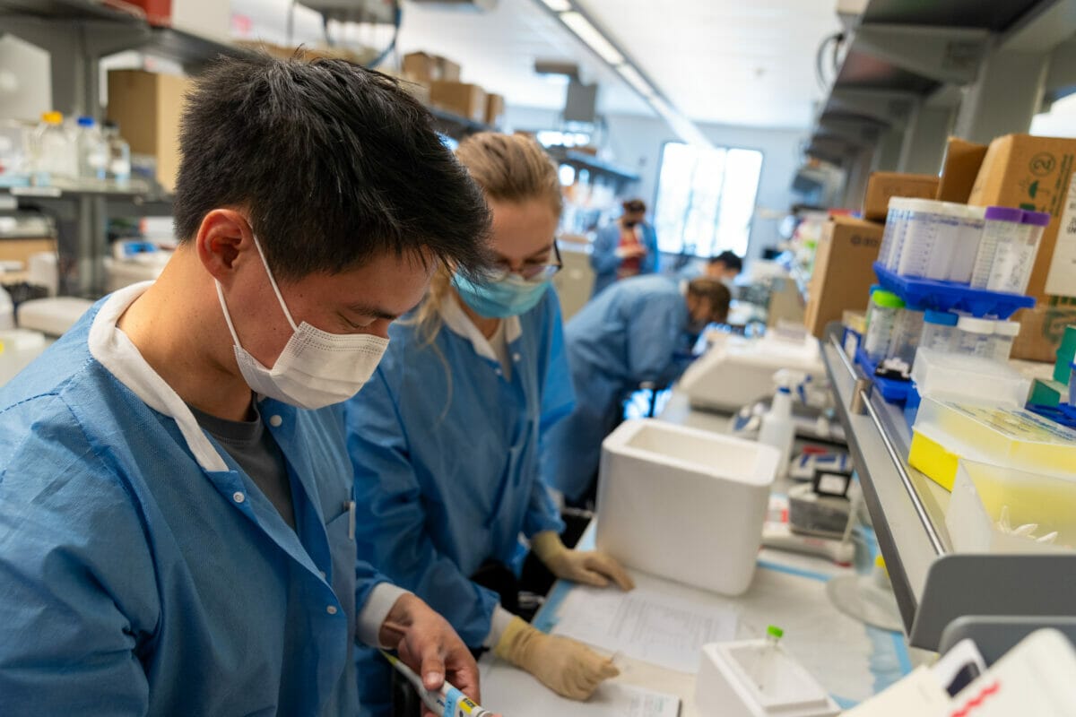 Two researchers in lab coats and face masks work in a laboratory filled with equipment and supplies.