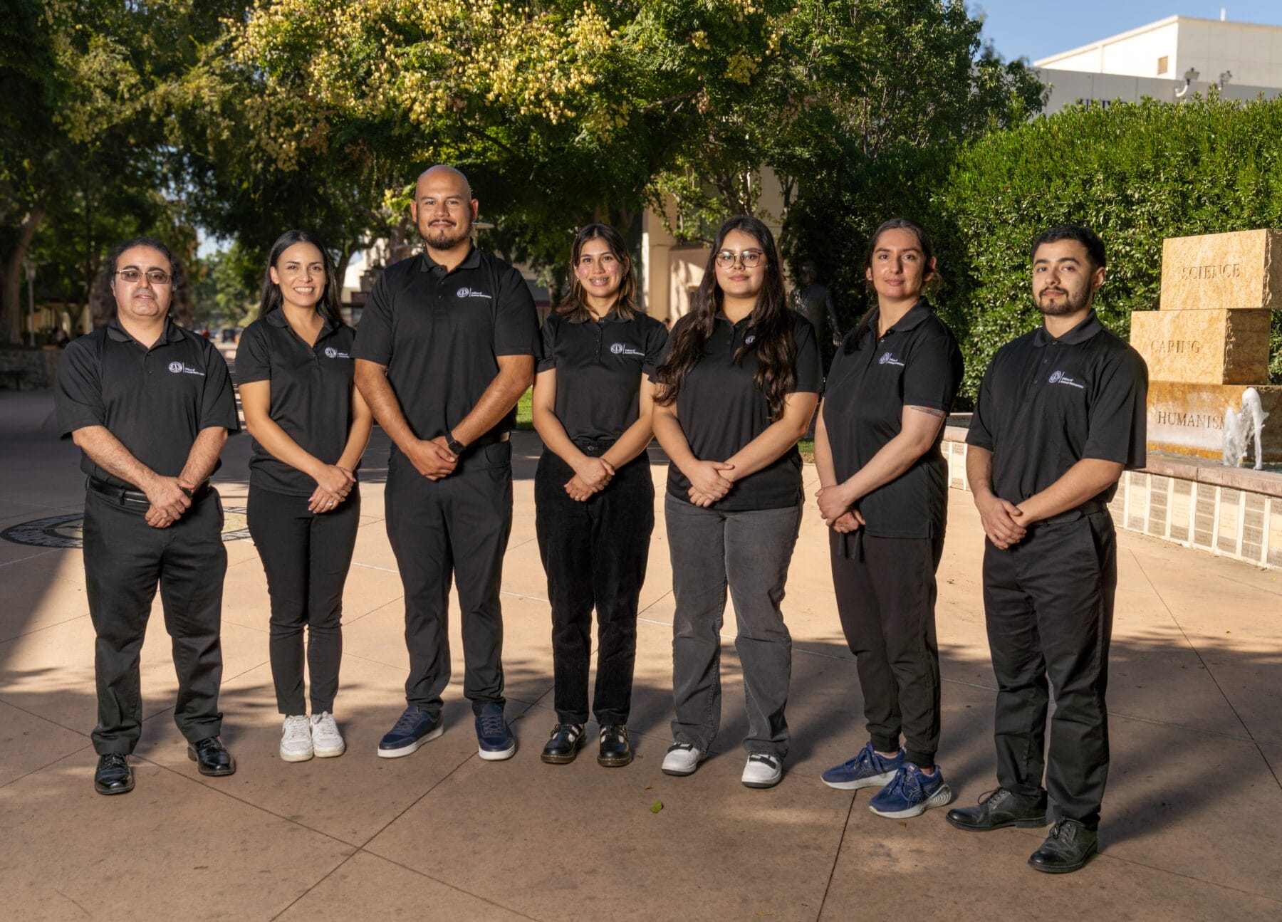 Seven people wearing black shirts and pants stand in a row outdoors on a sunny day, with trees and a stone fountain in the background.