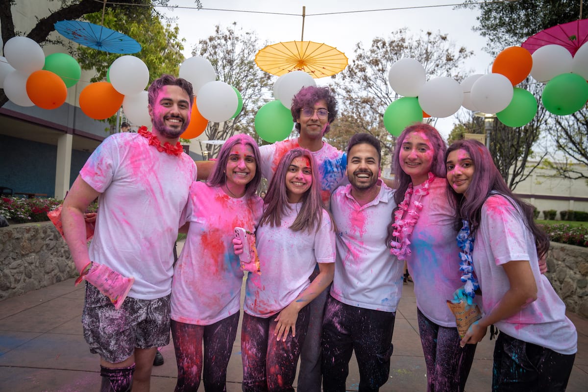 A group of people covered in colorful powder stand together, smiling at a festive outdoor event. They are wearing white shirts and leis, with colorful umbrellas and balloon strands in the background.