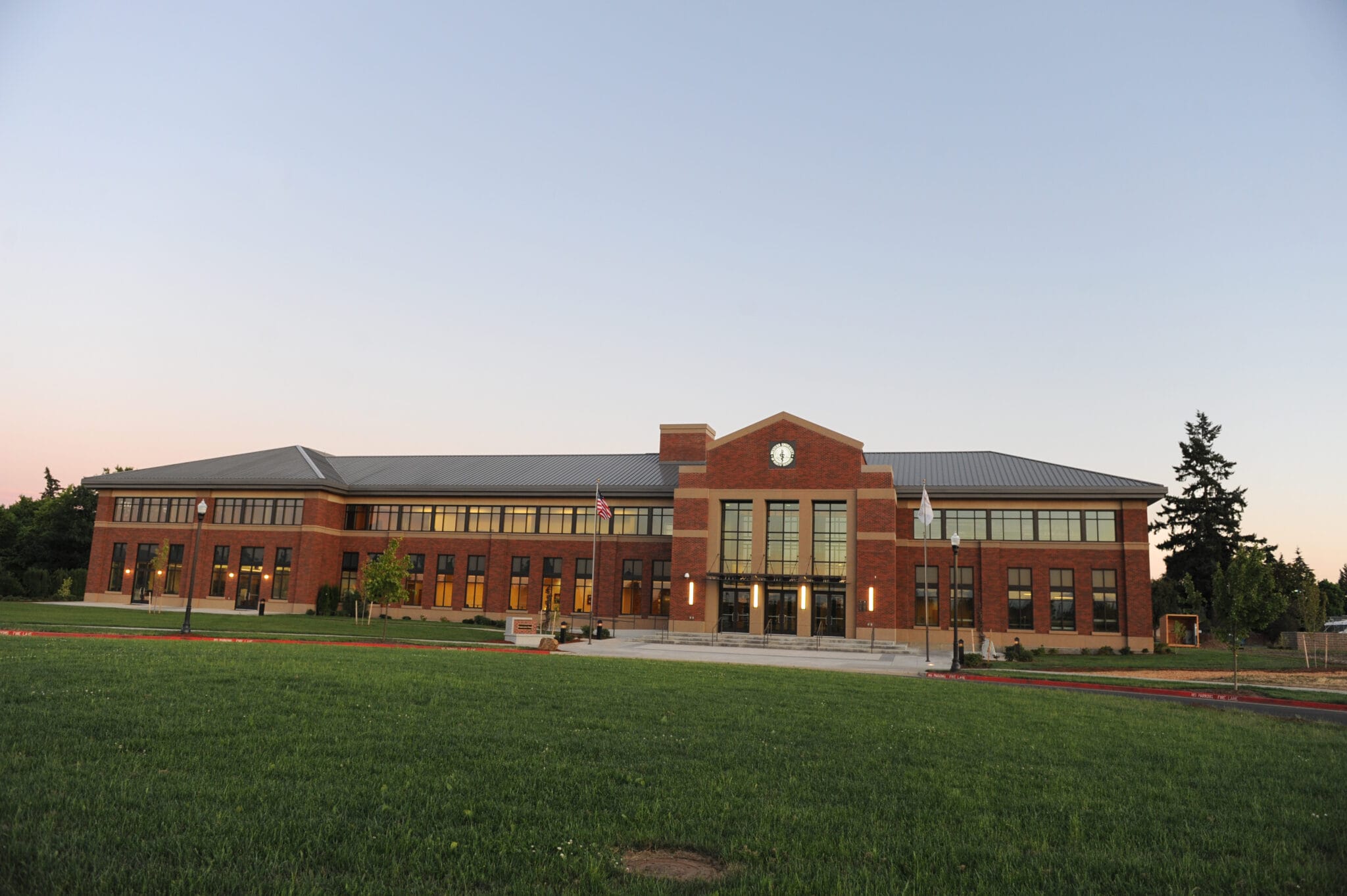 A large, two-story brick building with a clock above the entrance, surrounded by grass and trees, photographed at sunset.