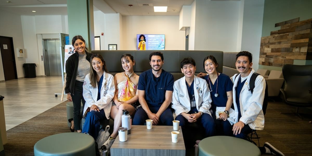 A group of seven colleagues, some in medical attire, sit and stand around a table in a lounge area with coffee cups and bags nearby.