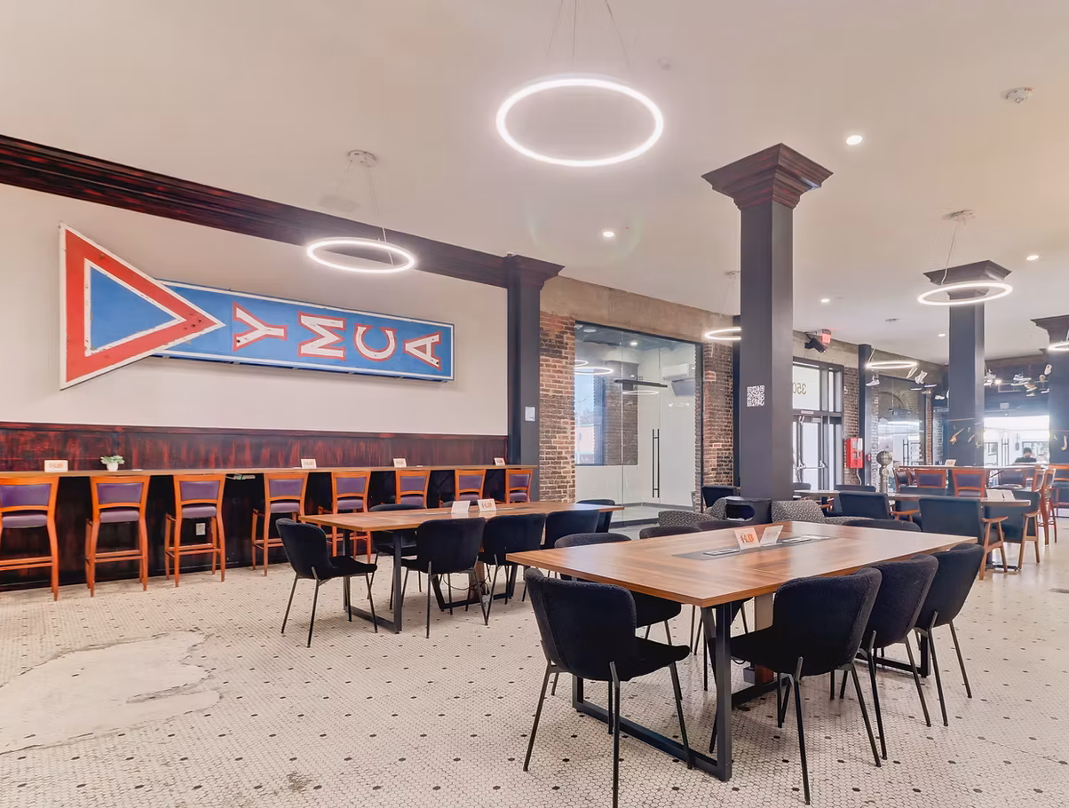 Modern dining area with black chairs and wooden tables, tiled floor, circular ceiling lights, and a large YMCA sign on the wall.