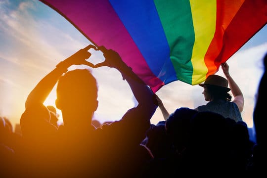 A crowd gathers under a large rainbow flag at sunset, with one person forming a heart shape with their hands in the foreground.
