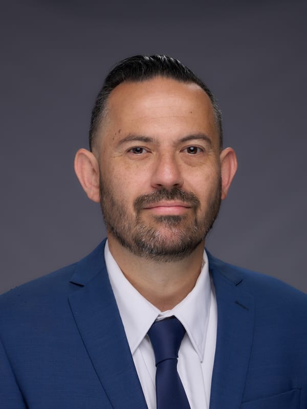 Professional head-and-shoulders portrait of a man in a blue suit and navy tie against a gray backdrop.
