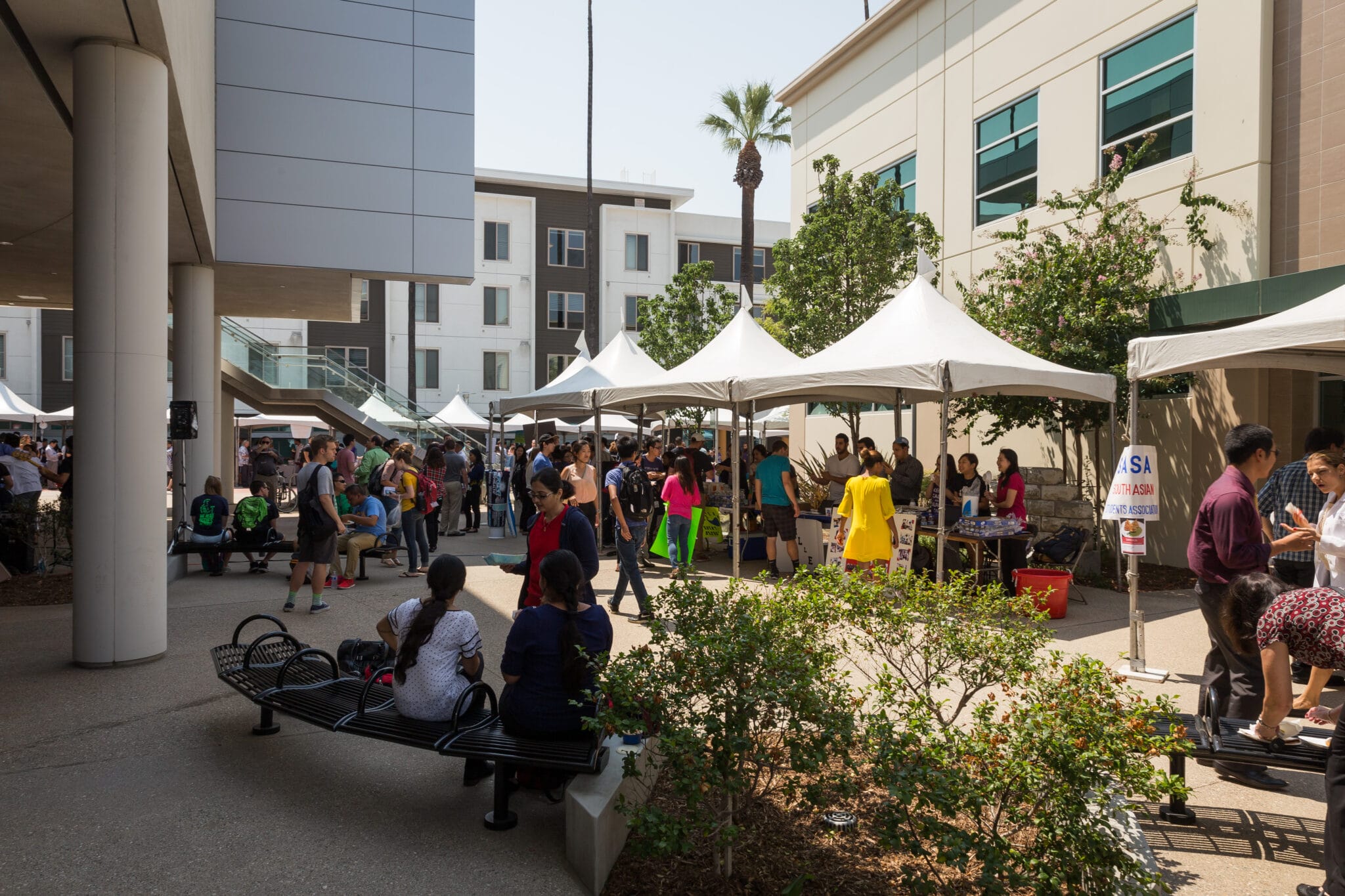 People attend an outdoor event with vendor tents set up between buildings; some are seated on benches while others visit booths.