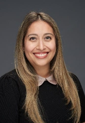 A woman with long, straight, light brown hair and a black top with a pink collar smiles at the camera against a plain gray background.