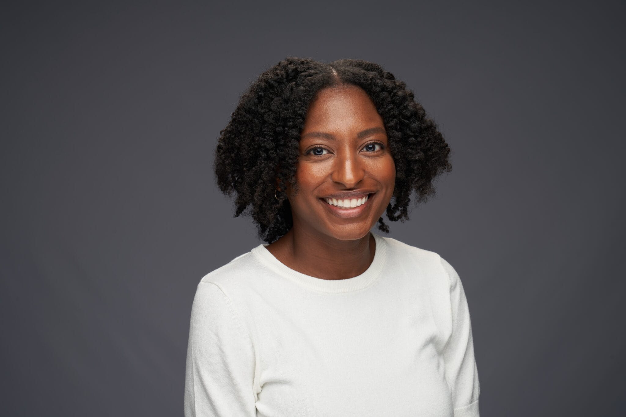 A woman with natural curly hair wearing a white top smiles at the camera against a plain gray background.