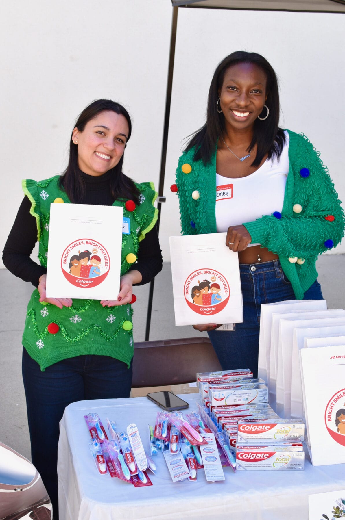 Two women stand behind a table with toothpaste and toothbrushes, holding up packets labeled with a cartoon dental care logo.