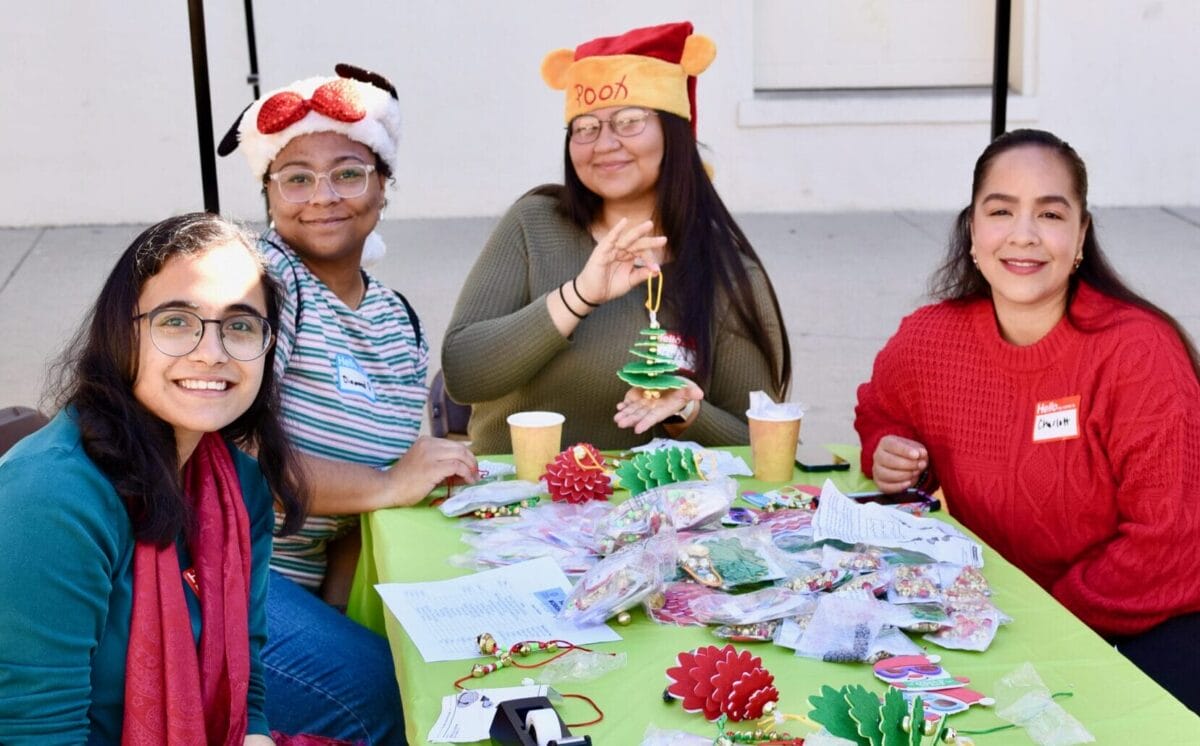 Four women sit at a table covered with Christmas crafts and decorations. Two of them wear festive hats, and all are smiling at the camera.