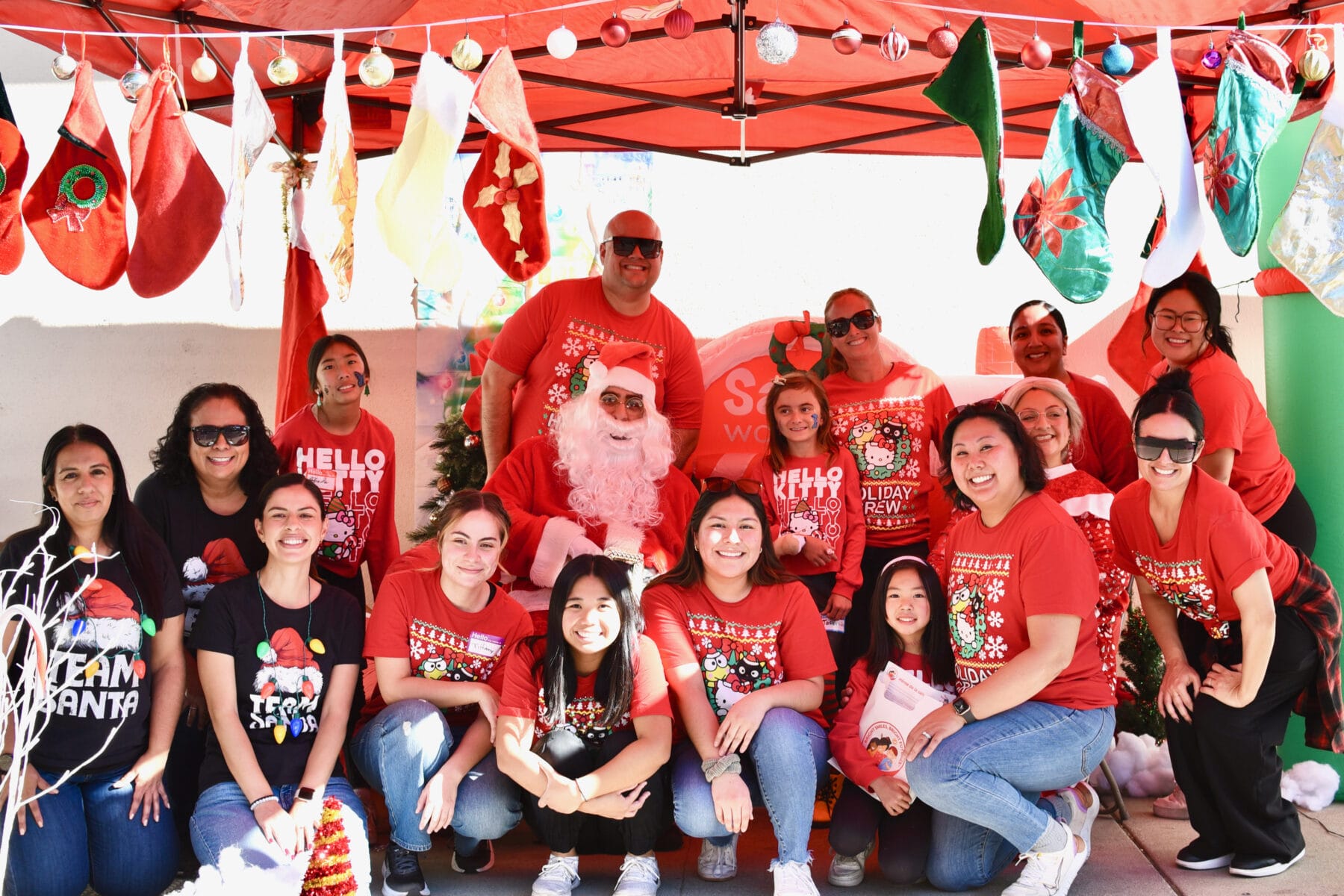 A group of people in festive attire pose with Santa Claus under a red tent decorated with stockings and ornaments.
