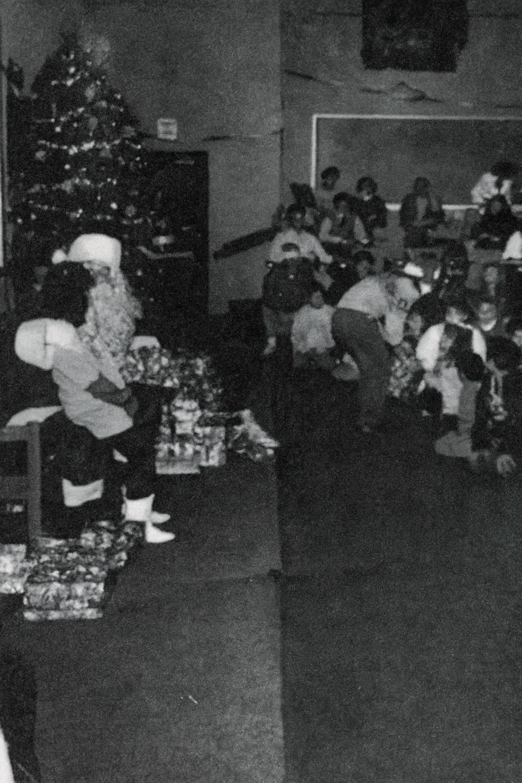 A person dressed as Santa Claus sits near a decorated Christmas tree and wrapped gifts while a group of people gather in a dimly lit room.