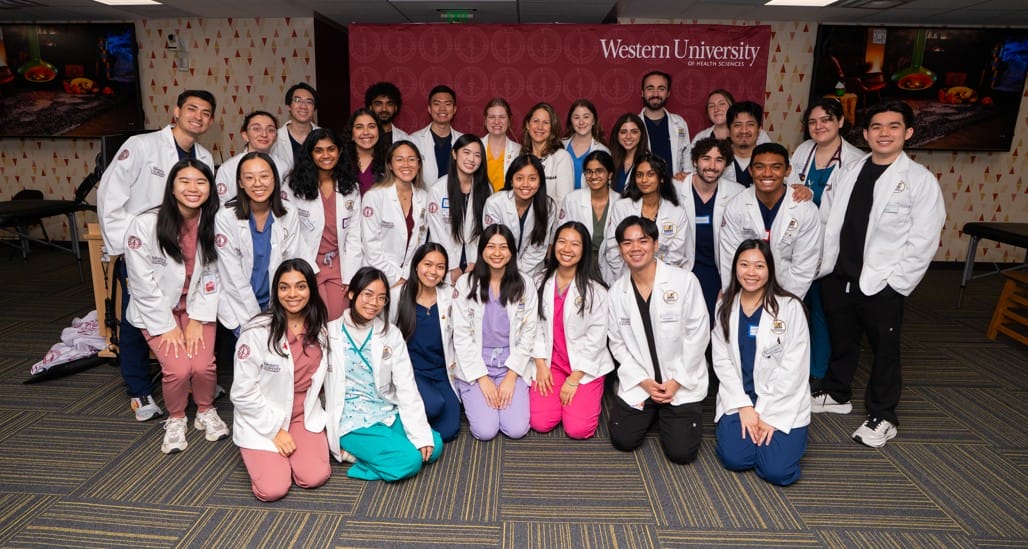 A group of students in white coats poses together in a classroom, with a “Western University” banner in the background.