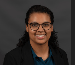 A person with curly dark hair, wearing glasses, a dark blazer, and a teal shirt, smiles against a plain dark background.