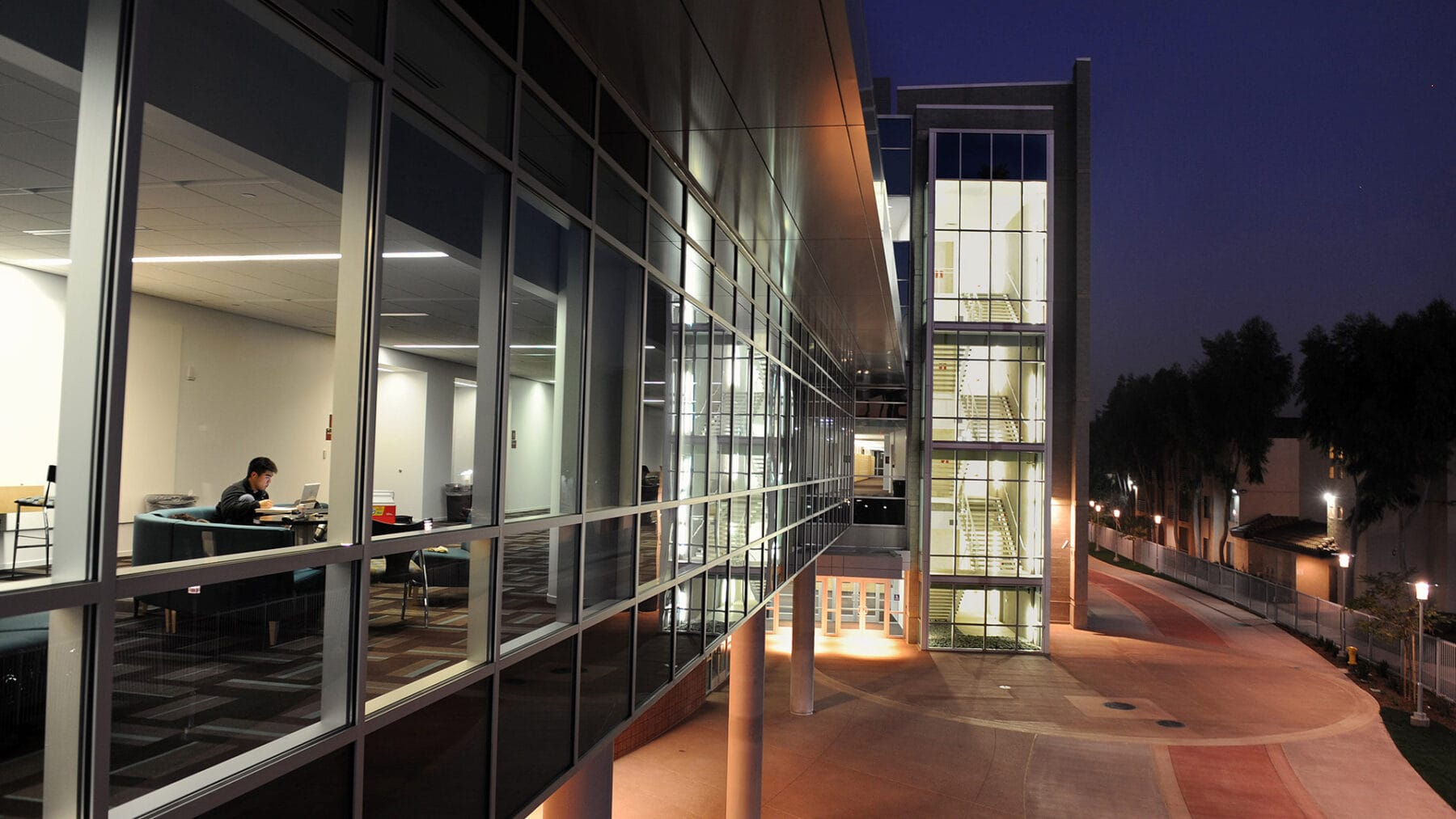 A person works at a desk inside a modern glass building at night; the building's lit stairwell and exterior walkway are visible.
