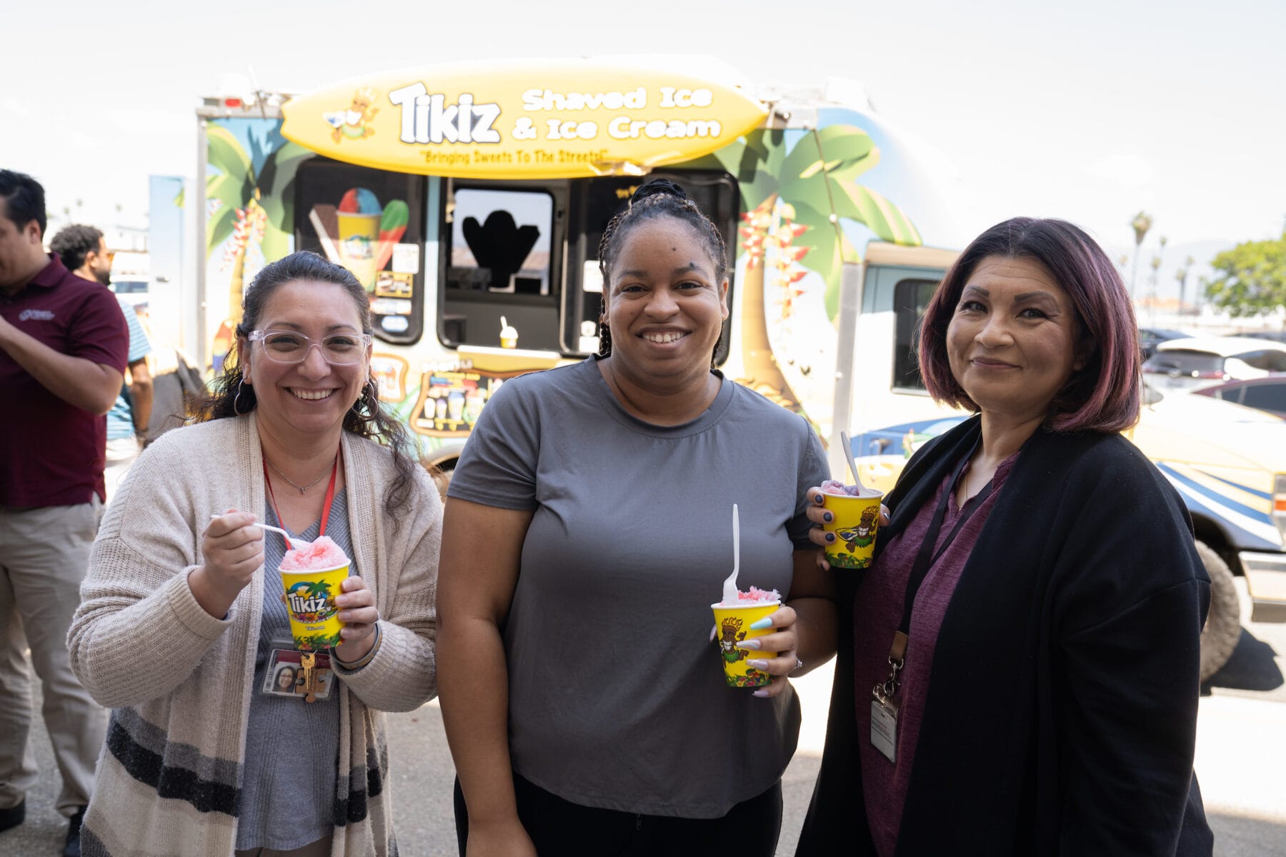 Three women stand outside holding cups of shaved ice in front of a Tikiz Shaved Ice & Ice Cream truck.