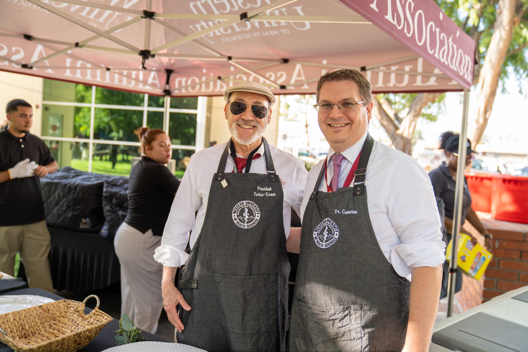 Two men wearing aprons stand side by side and smile at an outdoor event, with other people preparing food in the background.