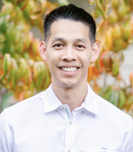 A man with short dark hair, wearing a white collared shirt, smiles at the camera with blurred green and orange foliage in the background.