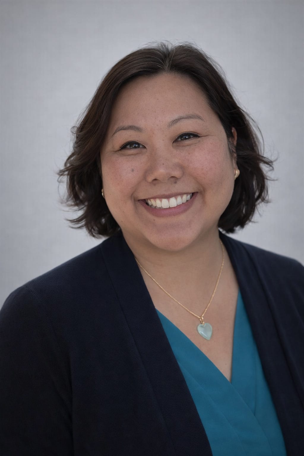 A woman with short brown hair, wearing a blue top, dark blazer, and a heart-shaped necklace, smiles at the camera against a plain light background.
