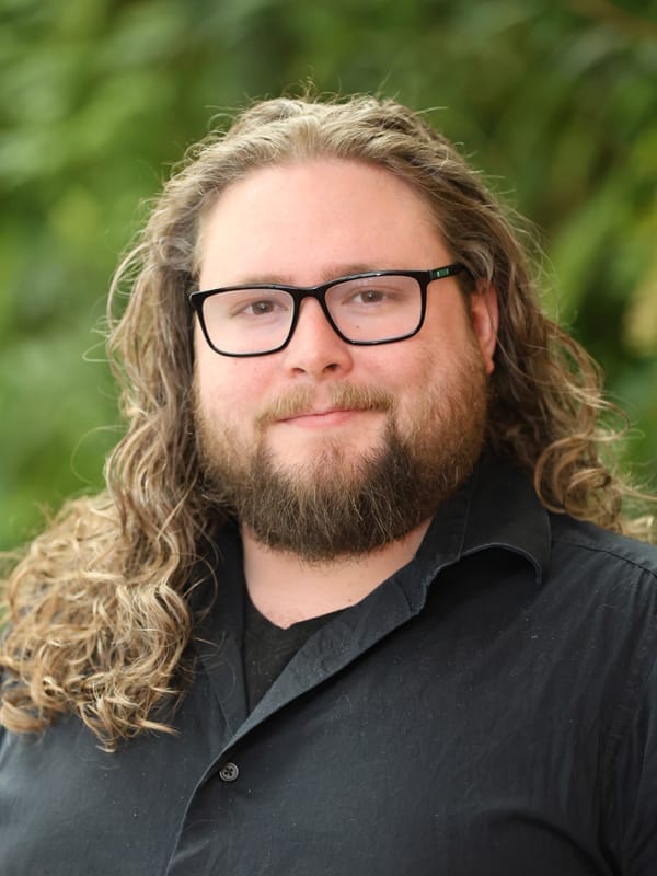 A person with long curly hair, a beard, and glasses is wearing a black shirt and standing outdoors in front of green foliage.