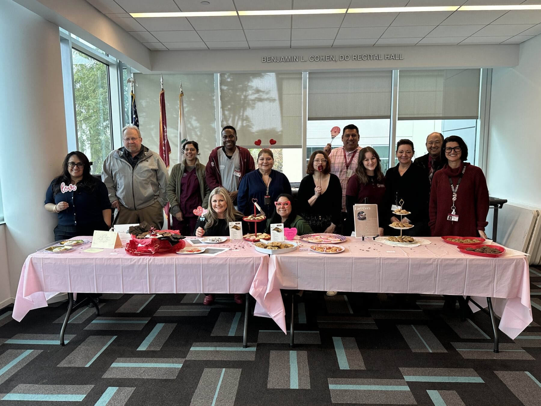 Thirteen people stand behind a table with pink decorations and treats in a brightly lit room, suggesting a festive or bake sale event.