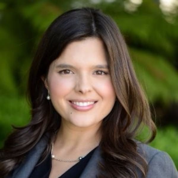 A professional headshot of a smiling woman with long brown hair, wearing a gray blazer and pearl earrings.