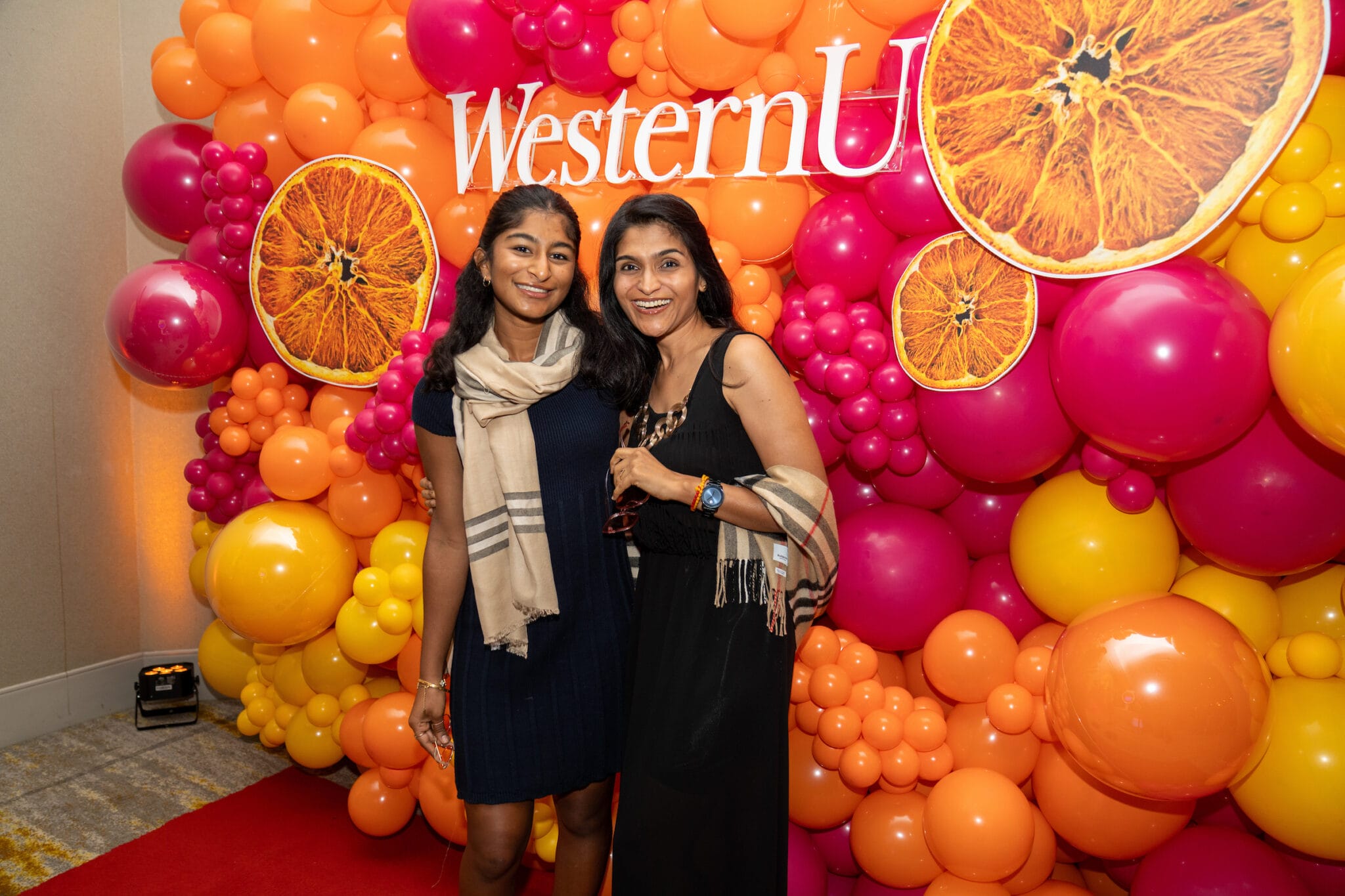 Two women stand smiling in front of a colorful balloon backdrop with orange slice decorations and a "WesternU" sign.