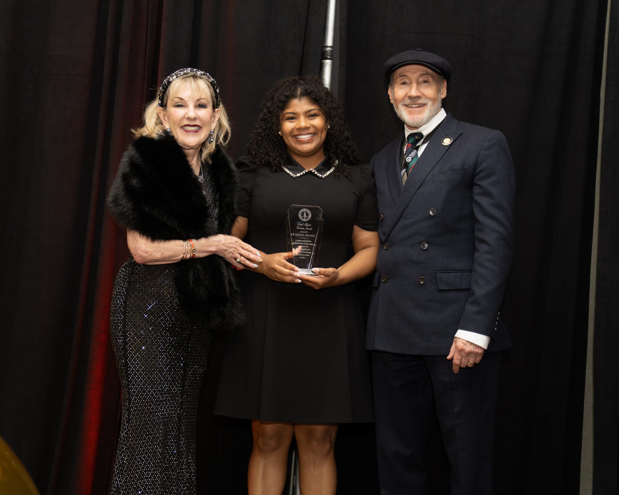 Three people stand together at an event; the woman in the center holds an award, while the others smile beside her against a black curtain backdrop.